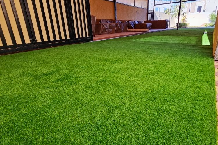 Covered area with artificial grass and patterned tiles, partially shaded by a wooden roof.