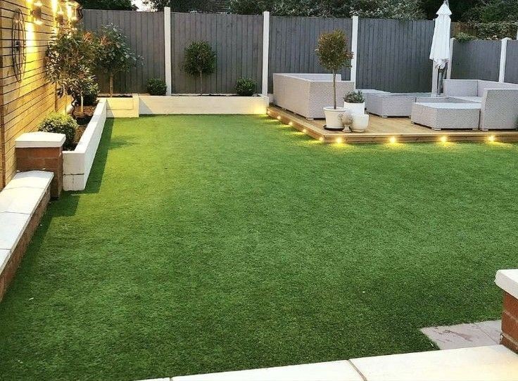 Modern backyard with artificial grass, garden plants, cozy seating area on wooden deck with ambient lighting.